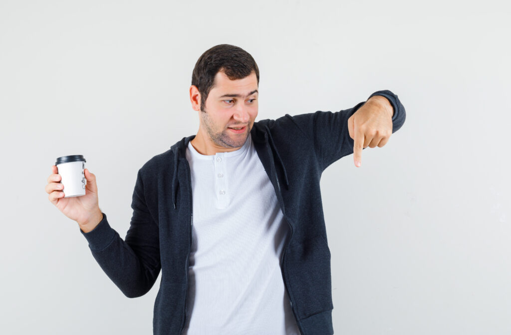young man in white t shirt and zip front black hoodie holding takeaway coffee cup and pointing down with index finger and looking surprised , front view.